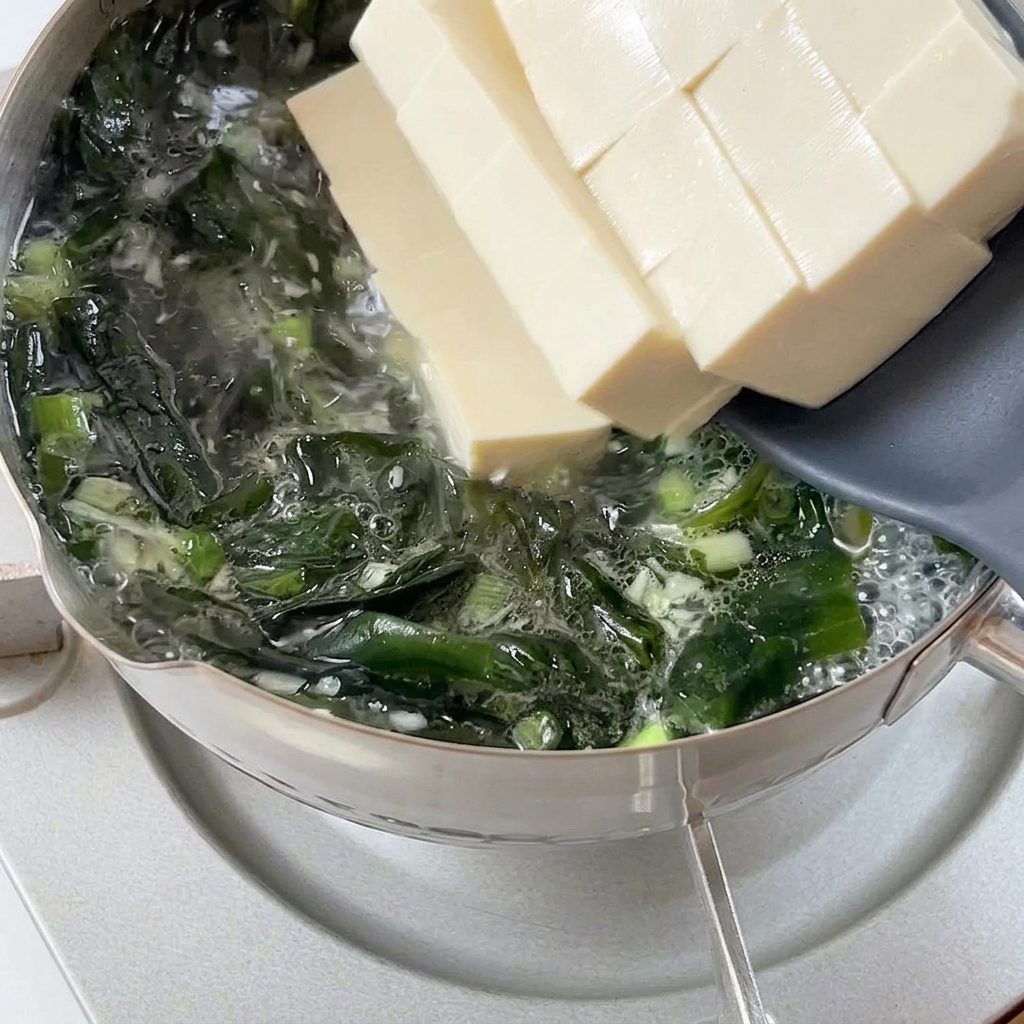 Grube kawałki białego, miękkiego tofu zsuwane z szarej łopatki do gotującego się bulionu.