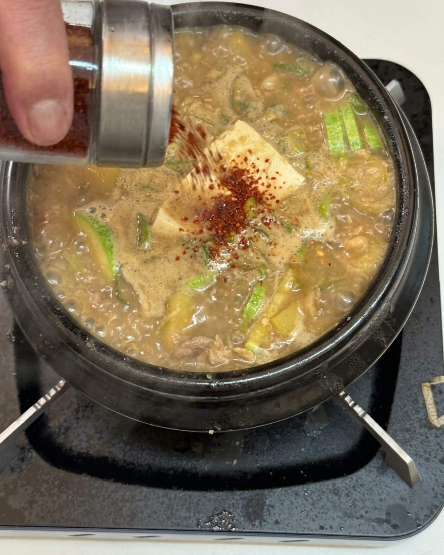 Czerwone płatki chili posypywane ze szklanego słoiczka na powierzchnię bulgoczącej zupy z tofu i cukinią.