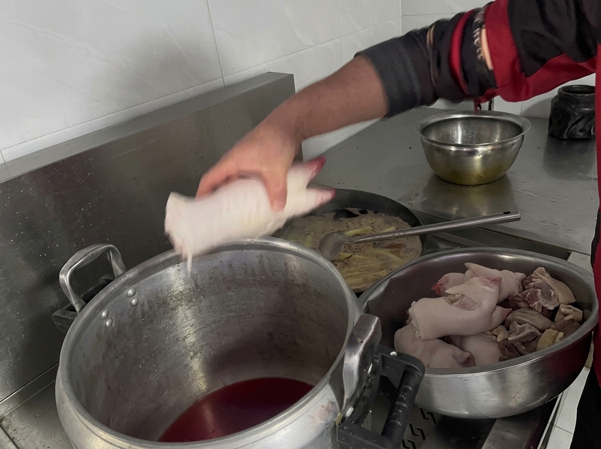 Dłoń wkładająca surowe nóżki wieprzowe do dużego, aluminiowego garnka wypełnionego ciemnym płynem.