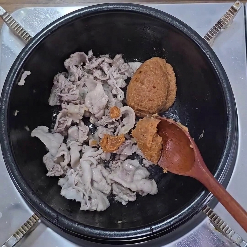 Łyżki brązowej pasty sojowej i pasty ssamjang dodawane do podsmażonego boczku w garnku.