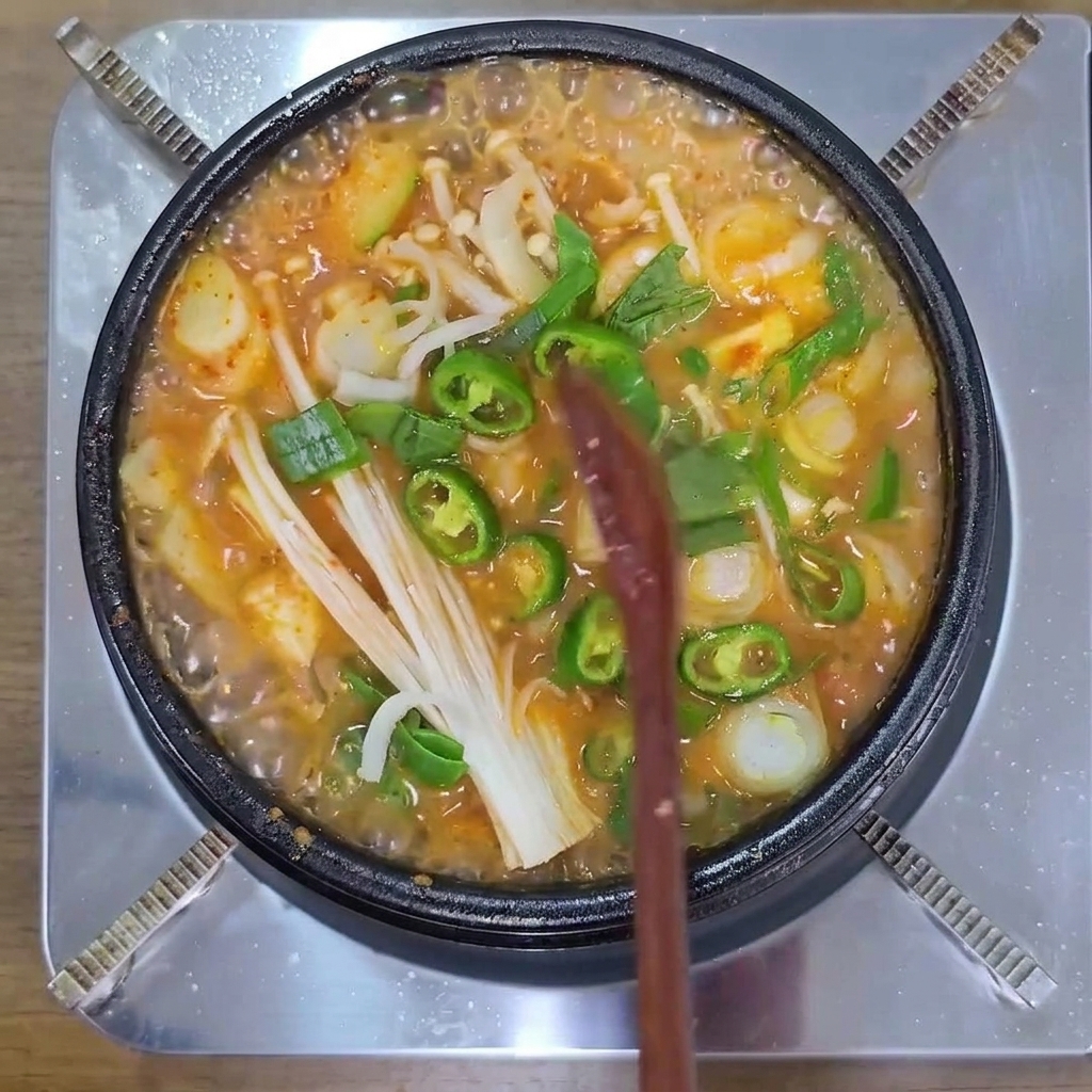 Plasterki zielonych papryczek chili, grzyby enoki i szczypiorek gotujące się w gorącym, bulgoczącym garnku z koreańską zupą.