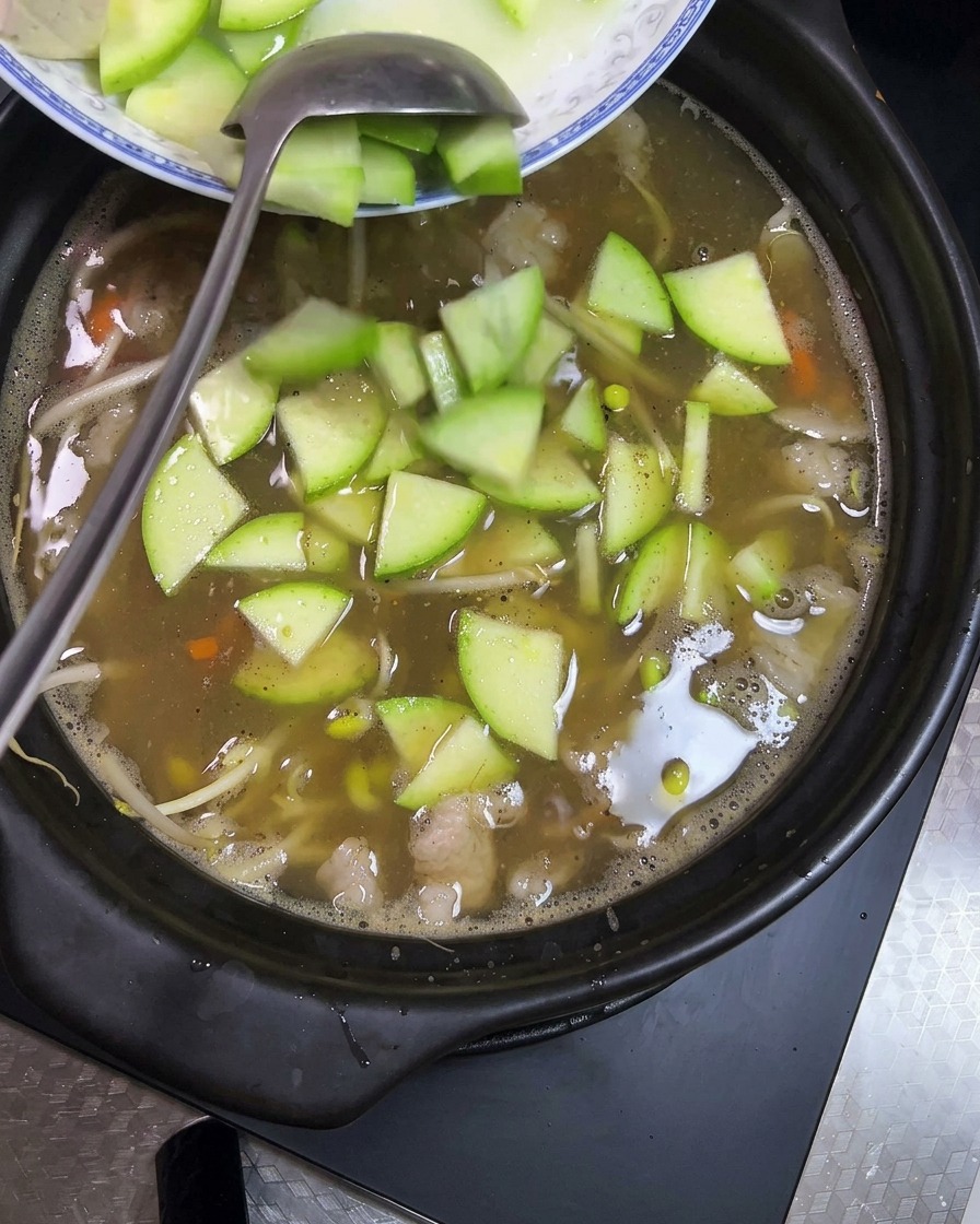 Kostki tofu i plasterki zielonej cukinii dodawane do glinianego naczynia z bulgoczącym gulaszem.