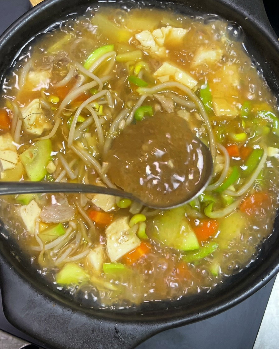 Metalowa łyżka z ciemną koreańską pastą mieszana z gotującym się w glinianym naczyniu gulaszem z tofu, cukinią, kiełkami i bulionem.
