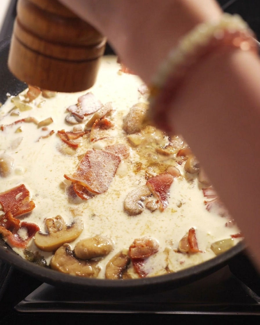 Ręka używająca drewnianego młynka do pieprzu nad patelnią z gotującym się sosem śmietanowym, boczkiem i pieczarkami.