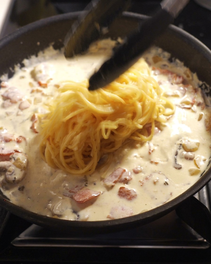Czarne szczypce kuchenne przekładające porcję ugotowanego spaghetti na patelnię z białym sosem, boczkiem i pieczarkami.