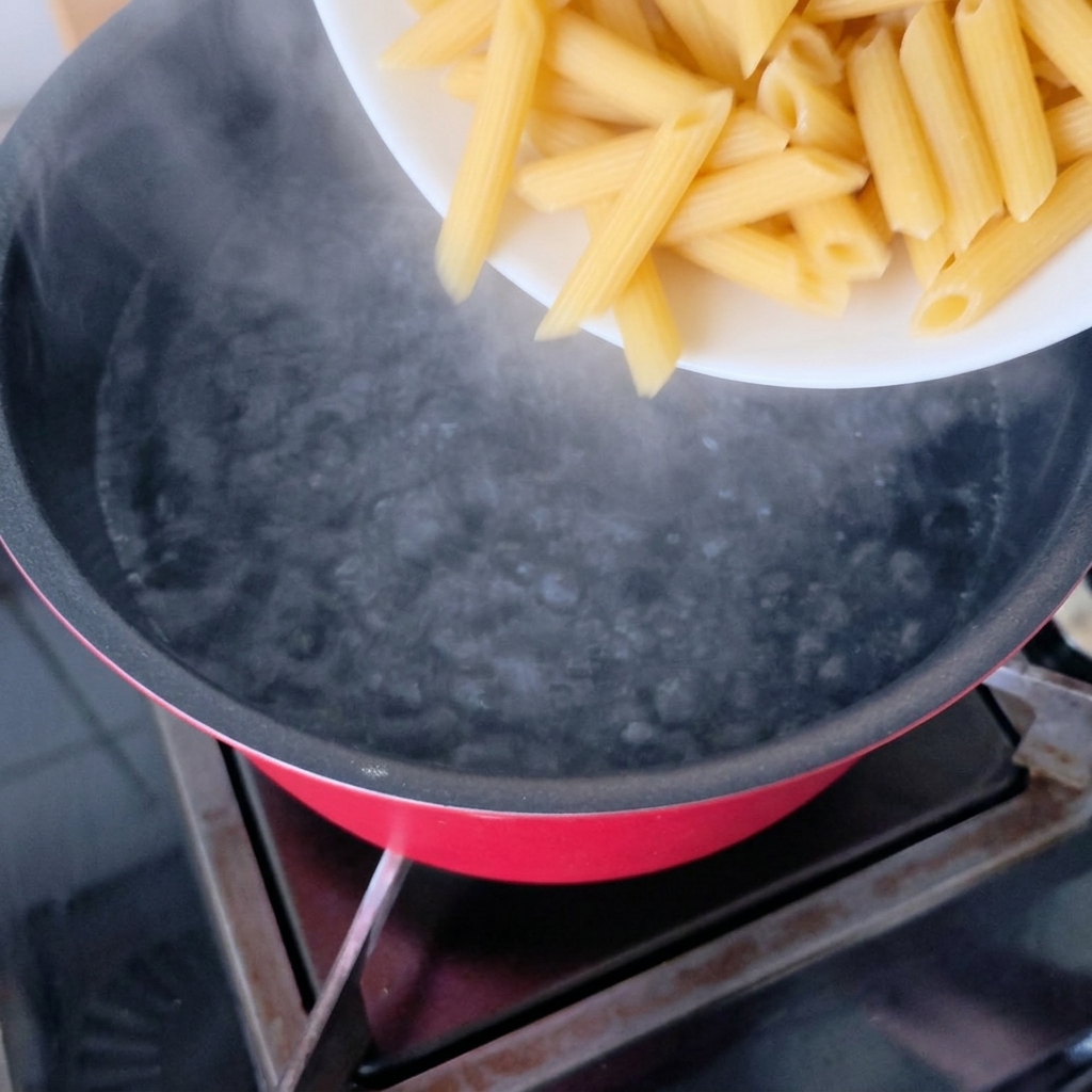 Suchy makaron penne wsypywany do garnka z wrzącą wodą.