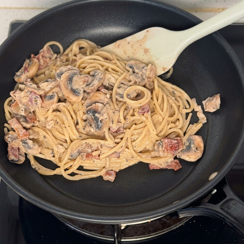 Patelnia wypełniona spaghetti w sosie z grzybami i boczkiem.
