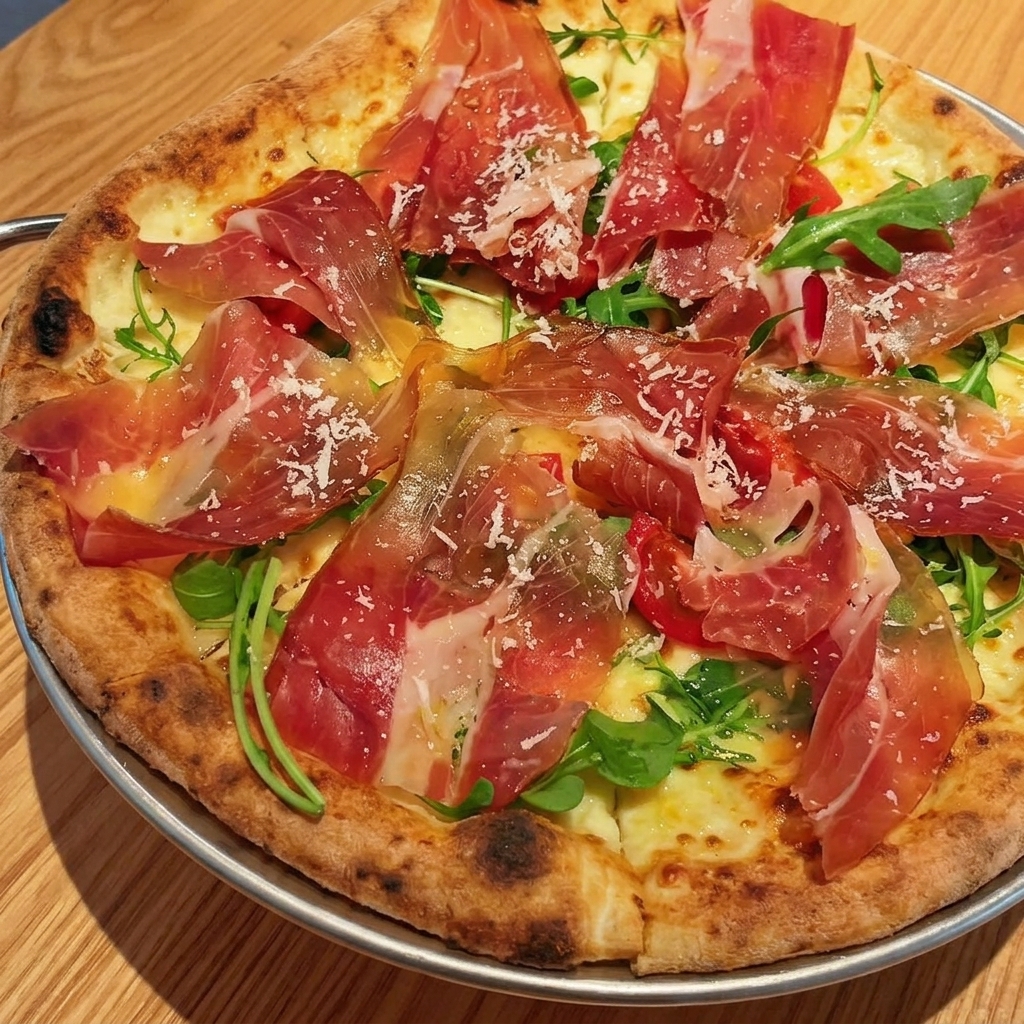 Cienkie, delikatne plastry surowej szynki parmeńskiej ułożone na świeżej rukoli i pomidorkach koktajlowych na upieczonej pizzy.