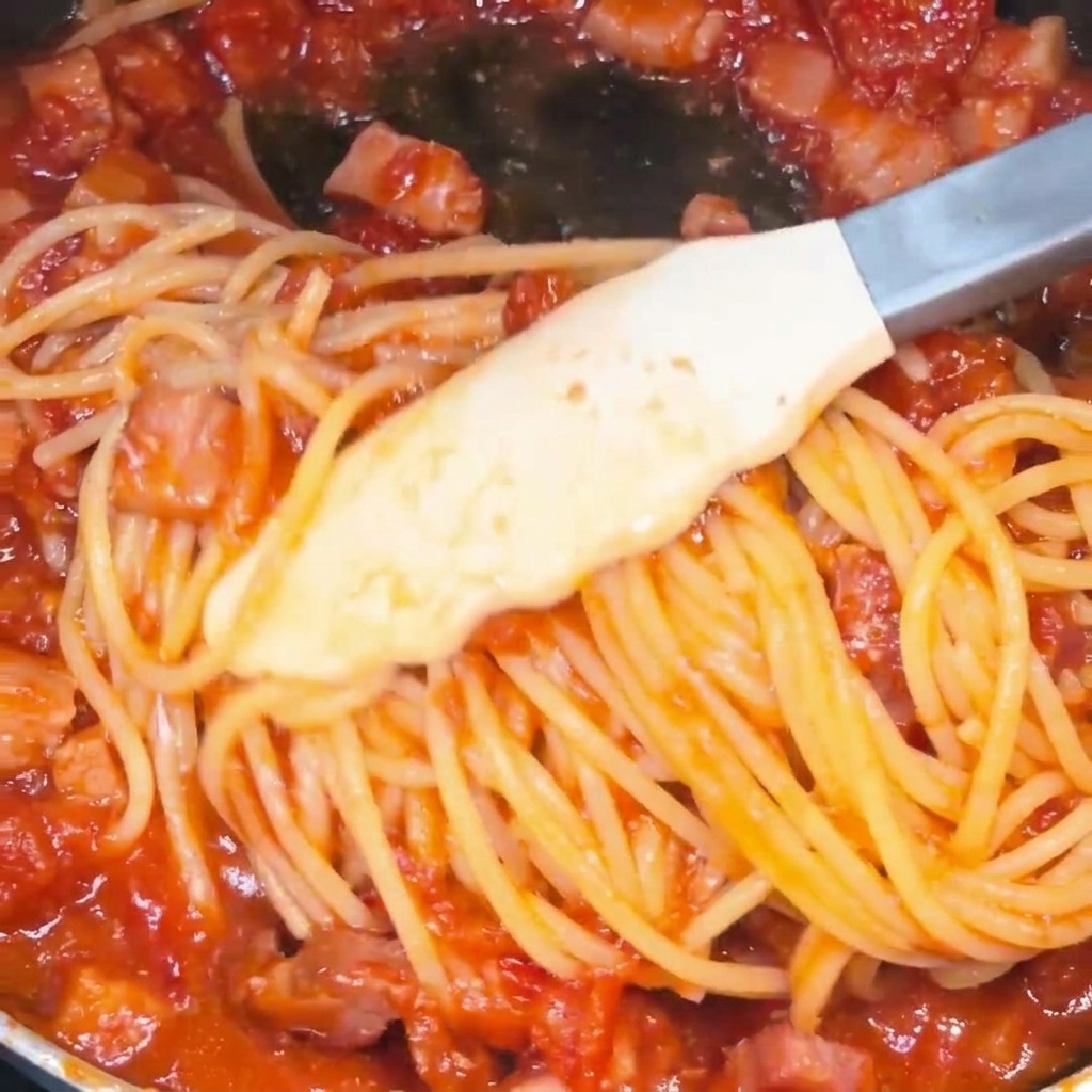 Spaghetti mieszane szczypcami na patelni, pokrywające się soczystym czerwonym sosem pomidorowo-boczkowym.