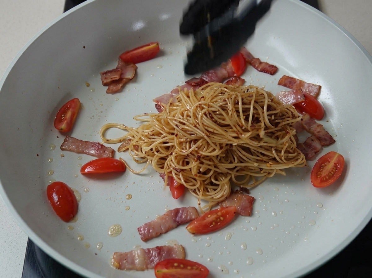 Doprawione spaghetti mieszane z boczkiem i pomidorami na patelni.