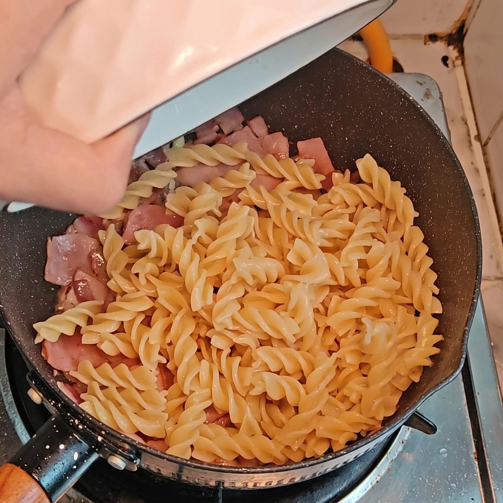 Makaron fusilli i kawałki szparagów dodawane na patelnię do boczku z cebulą.