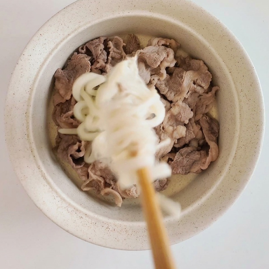 Skomponowana miska ze zblanszowanymi plastrami wołowiny, na których ułożony jest chłodzony makaron udon i grzyby enoki.