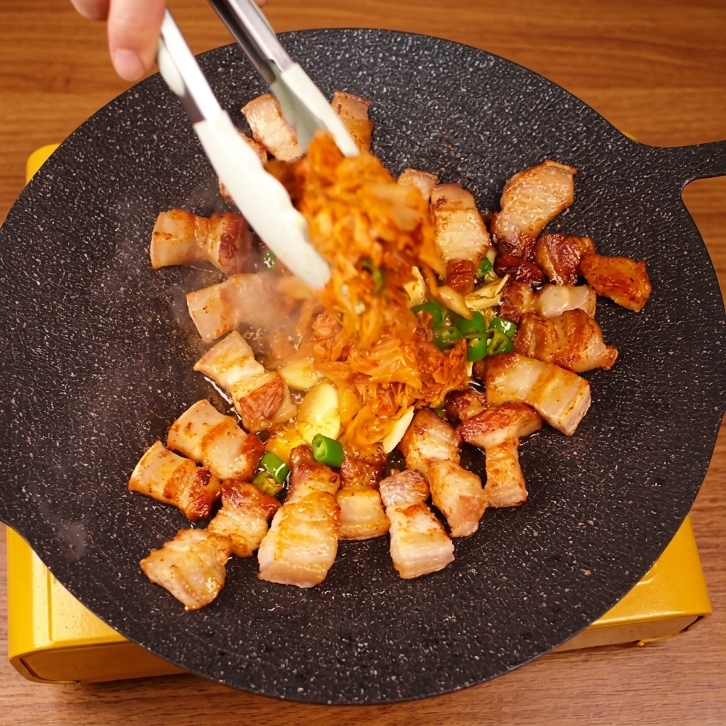 Dłoń za pomocą szczypiec miesza boczek z kimchi na gorącej patelni.