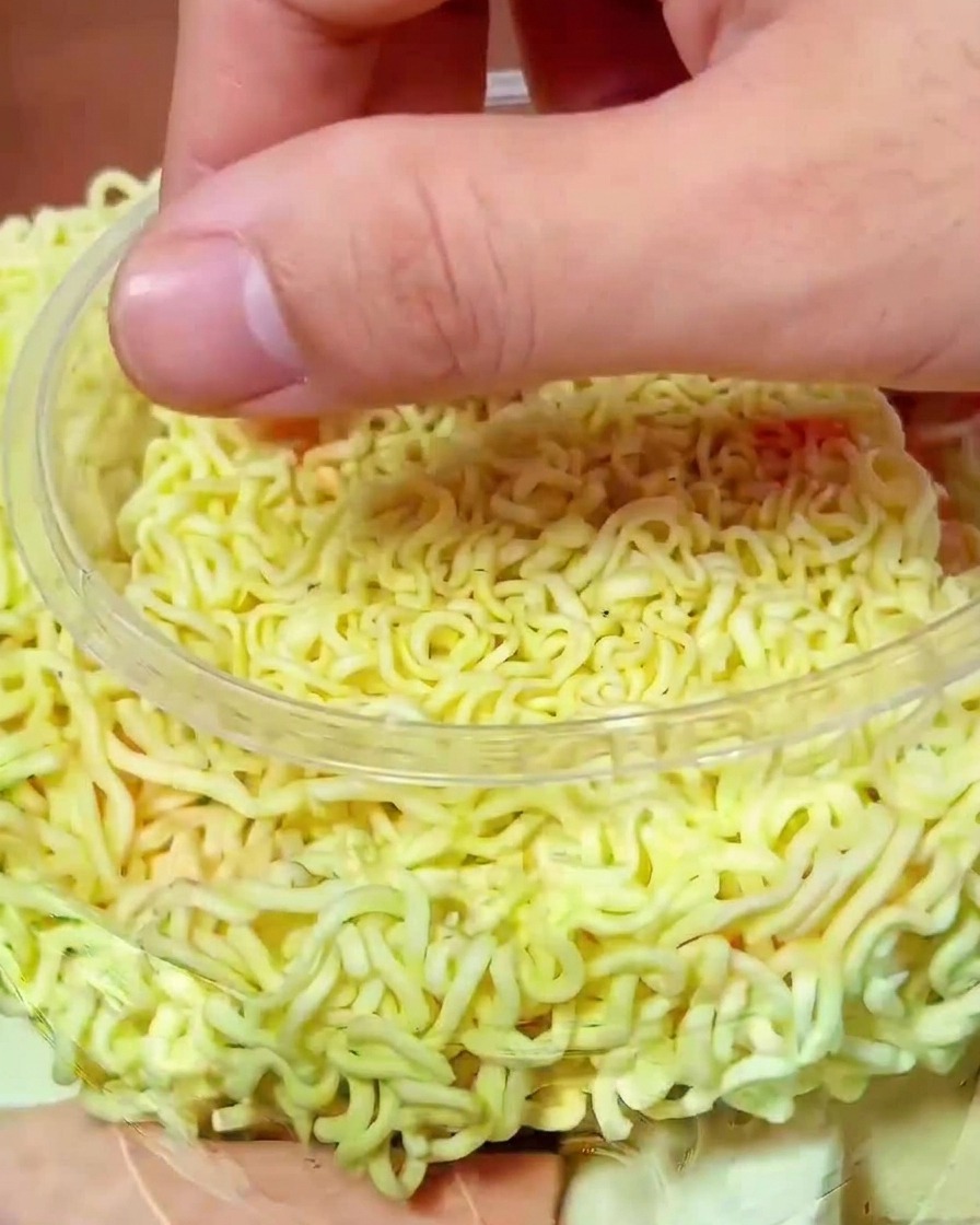 Dłoń układająca kawałki czerwonego kimchi na surowym makaronie w garnku.