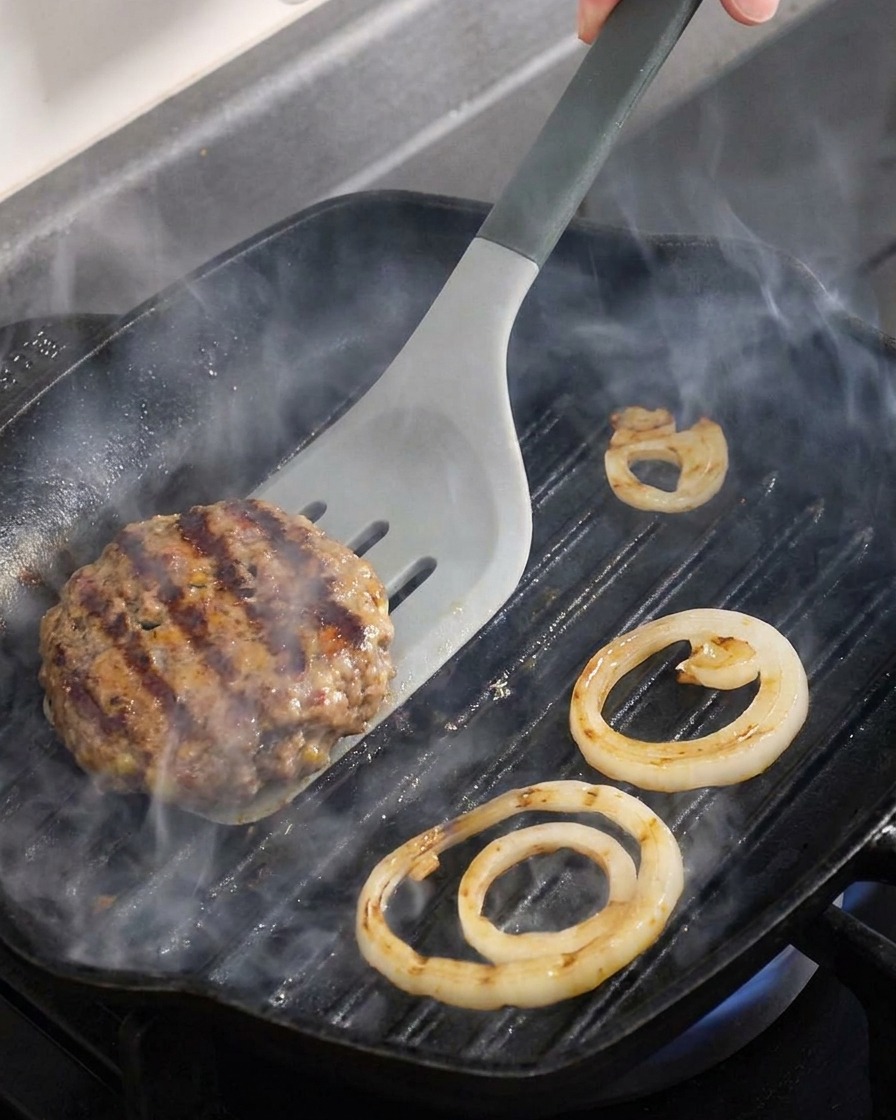 Zbliżenie na kotlet wołowy i krążki cebuli odwracane na patelni grillowej.
