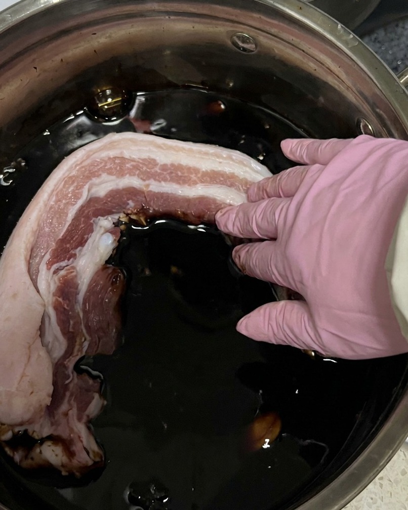 Paski surowego boczku zanurzane w garnku z ciemną marynatą sojową.