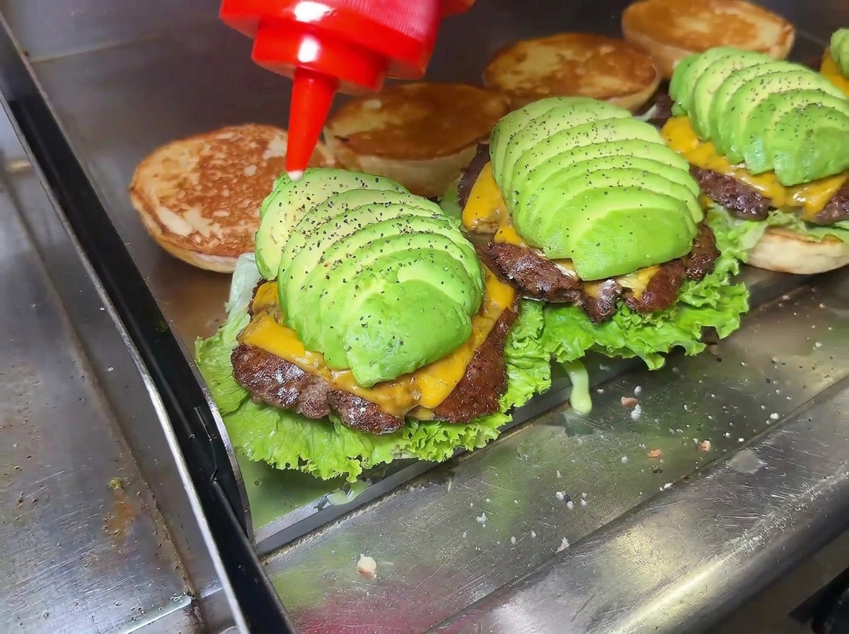 Zbliżenie na dłoń wyciskającą czerwoną butelkę sosu, polewającą idealnie ułożonego cheeseburgera z awokado na patelni.