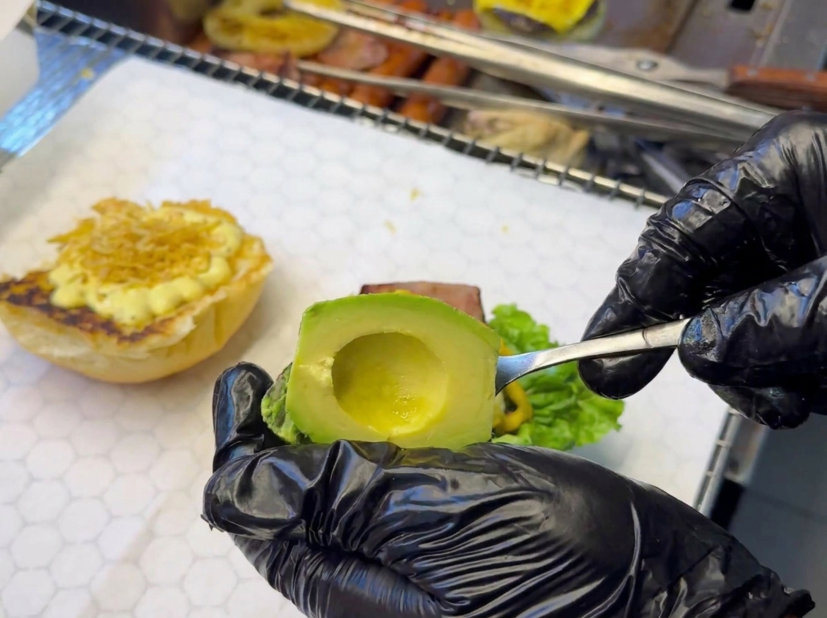 Dłoń w czarnej rękawiczce kładąca plasterki awokado na burgerze.