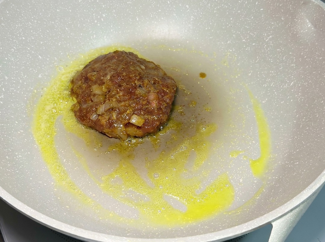 Surowy, doprawiony kotlet wołowy smażący się na jasnej patelni na małym ogniu.
