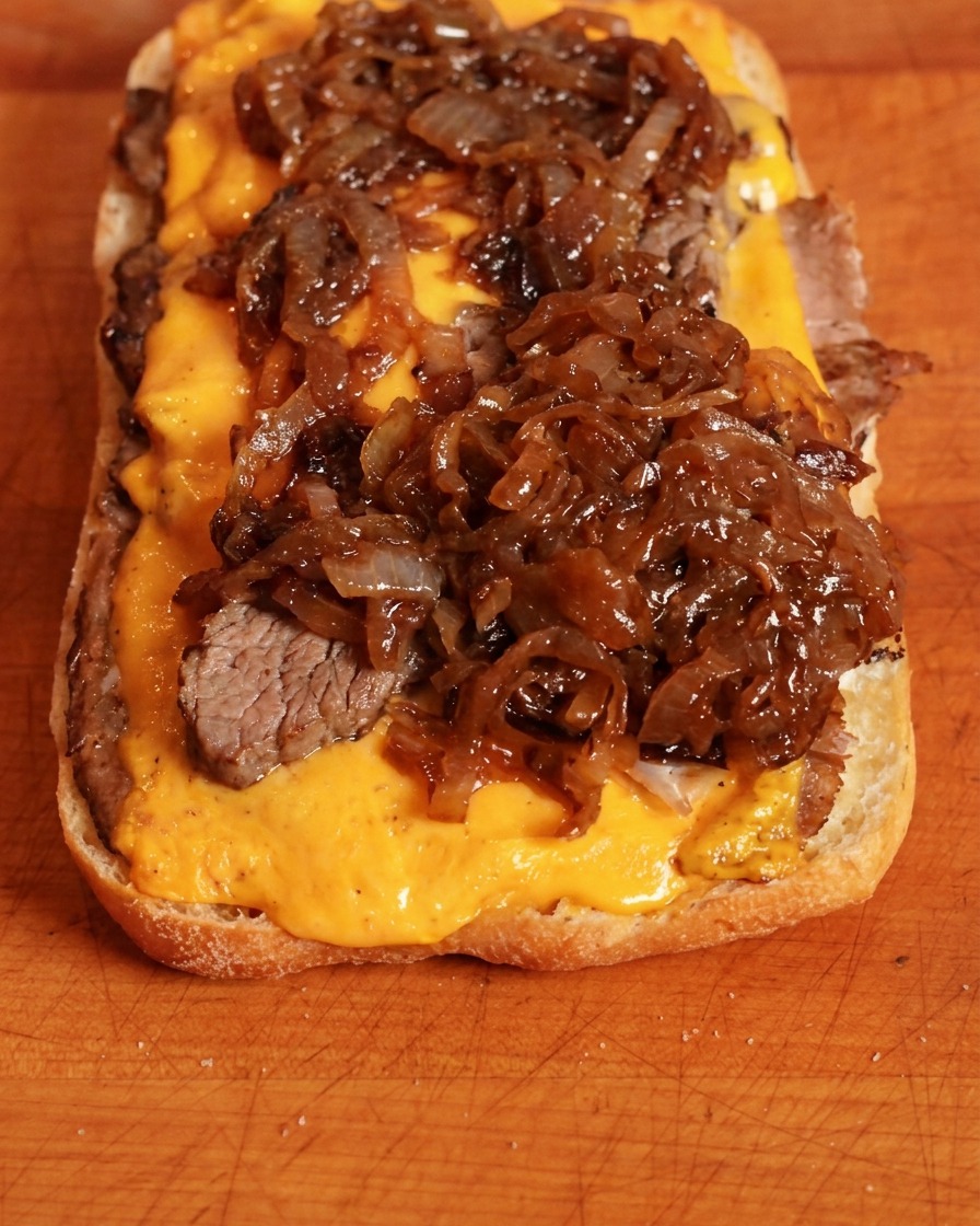 Ciemnobrązowa karmelizowana cebula nakładana łyżką na żółty ser cheddar na spodzie kanapki.