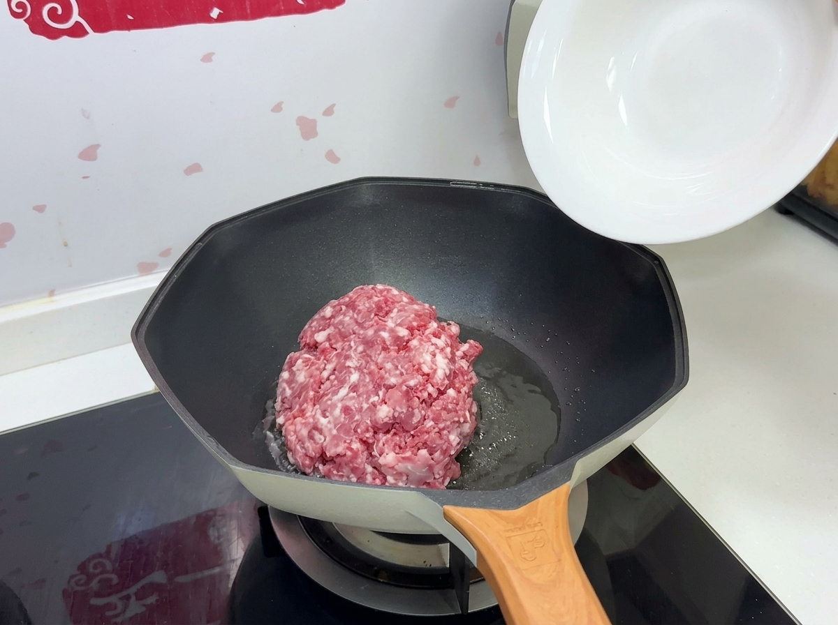 Kawałek surowego mięsa mielonego wrzucany z białej miski na rozgrzaną patelnię z olejem.