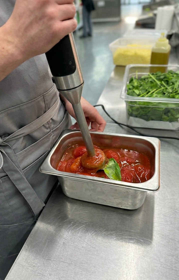 Zbliżenie na blender ręczny miksujący całe pomidory bez skóry i liście bazylii na gładki, jasnoczerwony sos do pizzy.