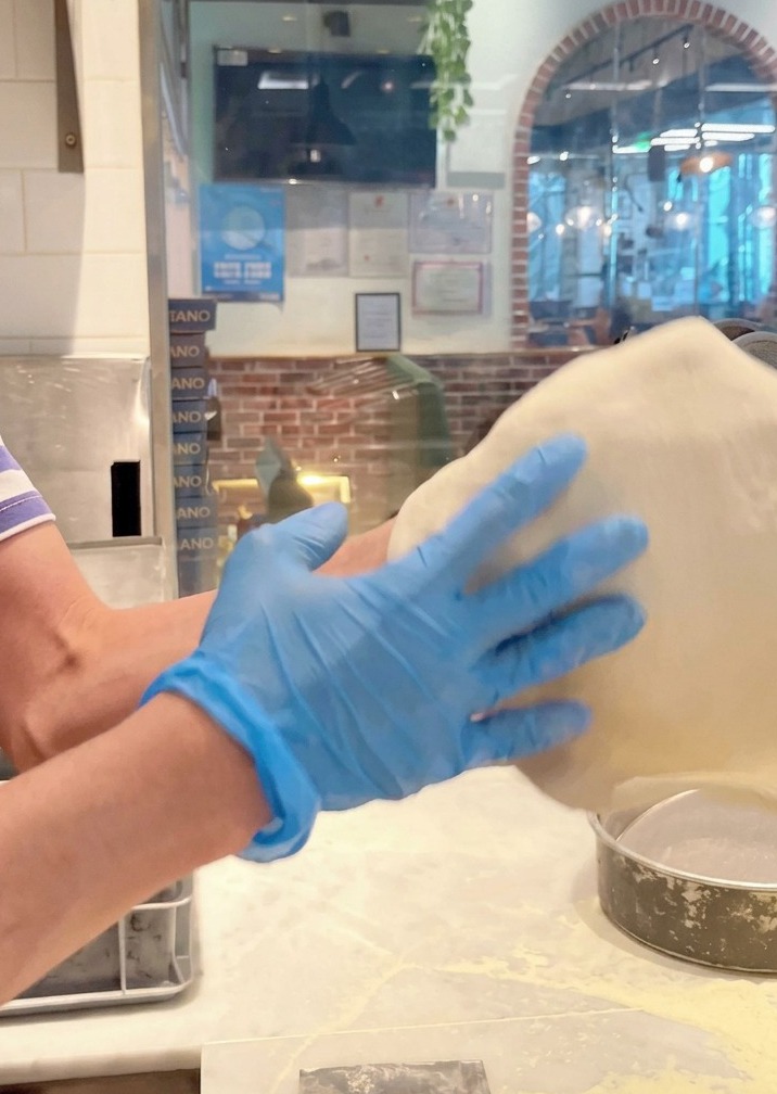 Ciasto na pizzę podrzucane i rozciągane w powietrzu przez dłonie w niebieskich rękawiczkach.