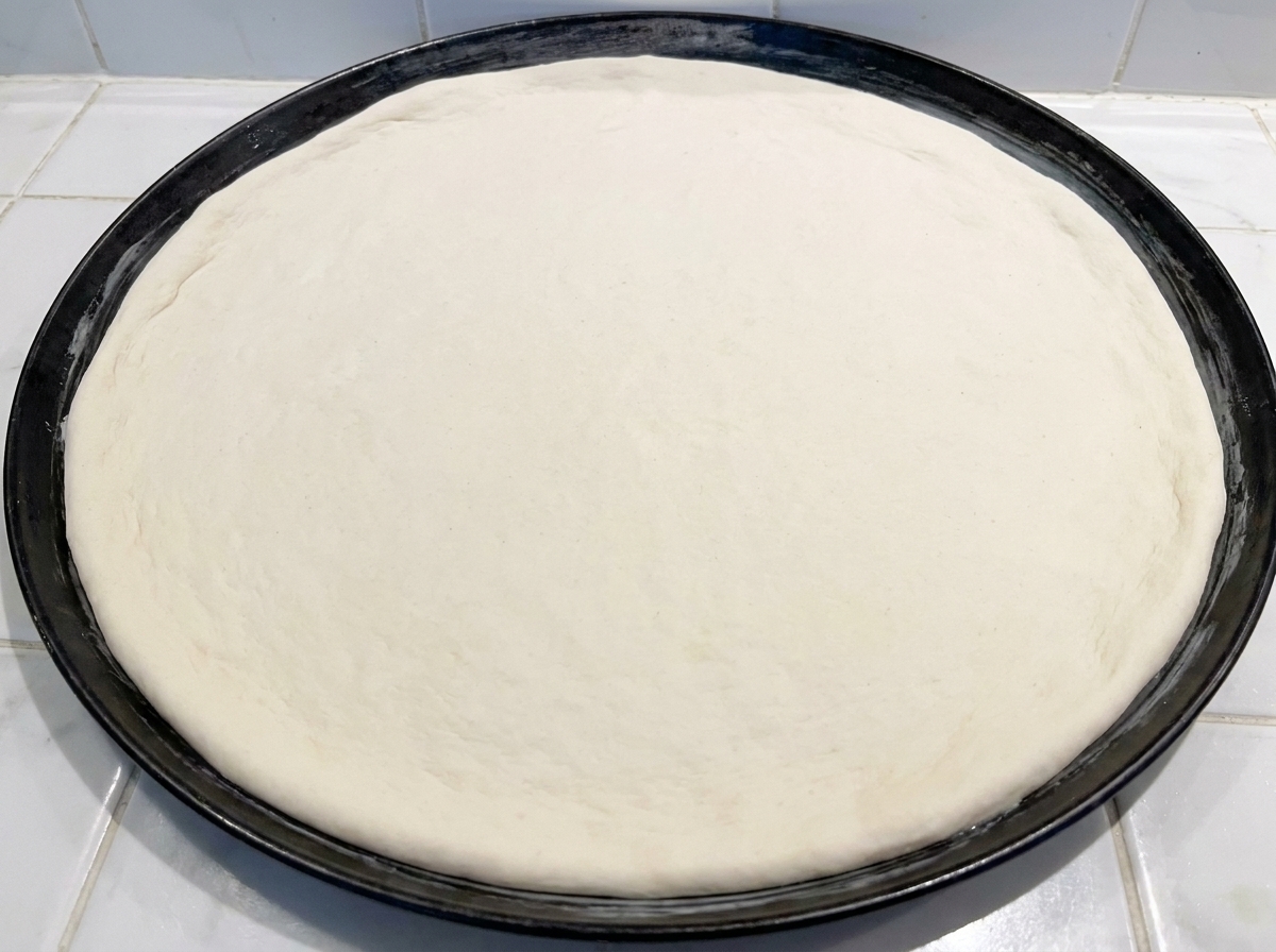Okrągłe, surowe ciasto na pizzę dociskane do czarnej, metalowej formy do pieczenia.
