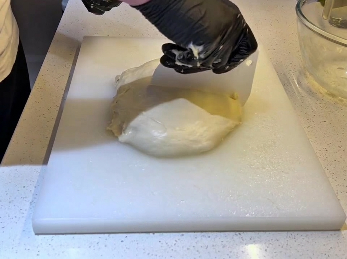 Dłonie w rękawiczkach używające plastikowej skrobki do podzielenia ciasta na pół.