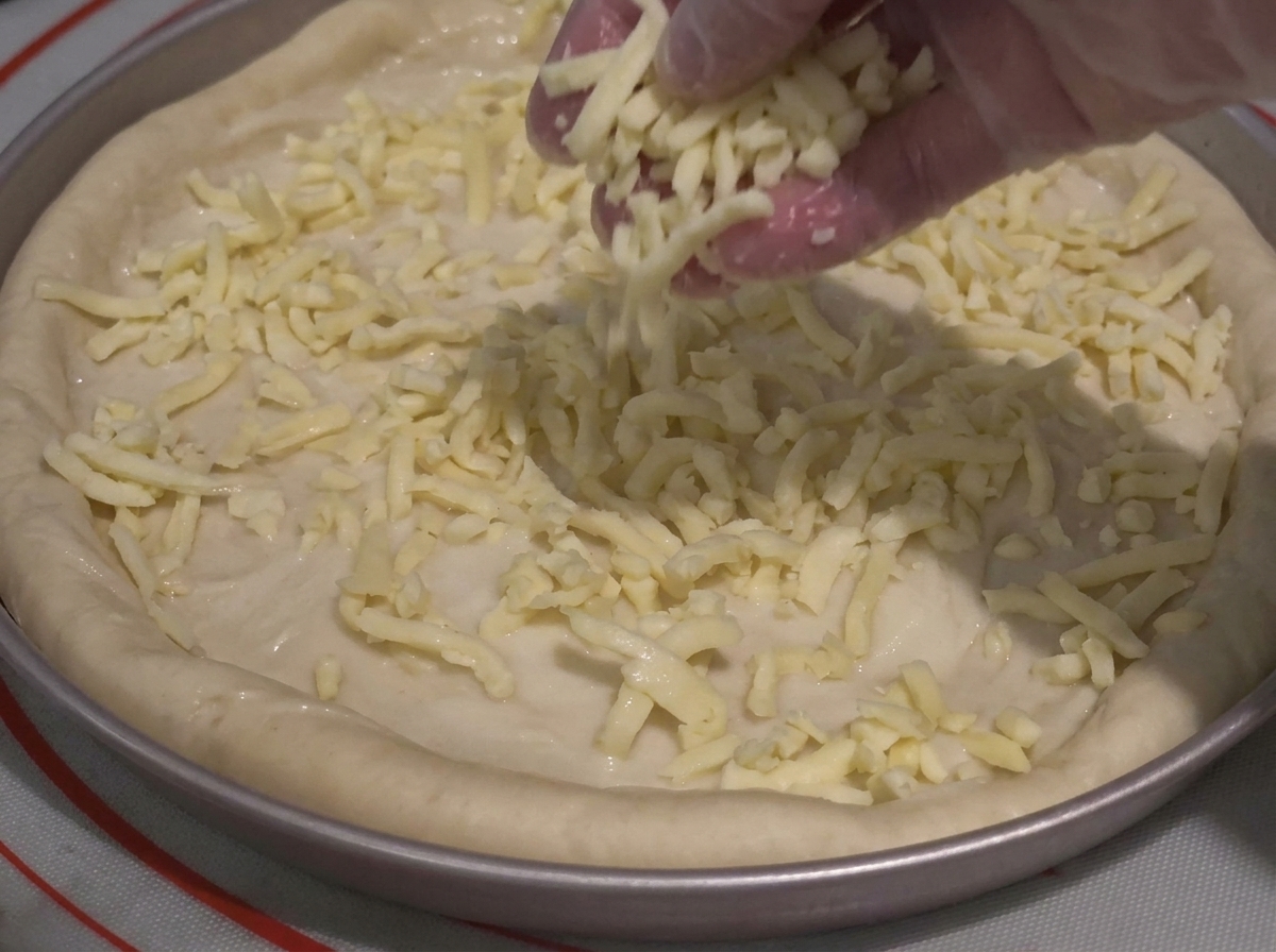 Ręka w rękawiczce hojnie posypująca startym serem ciasto do pizzy posmarowane sosem w blasze.
