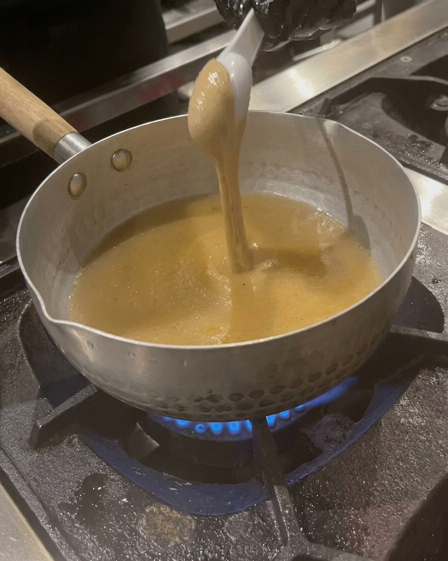 Łyżka gęstej, beżowej pasty sezamowej mieszana z bulionem w garnku.