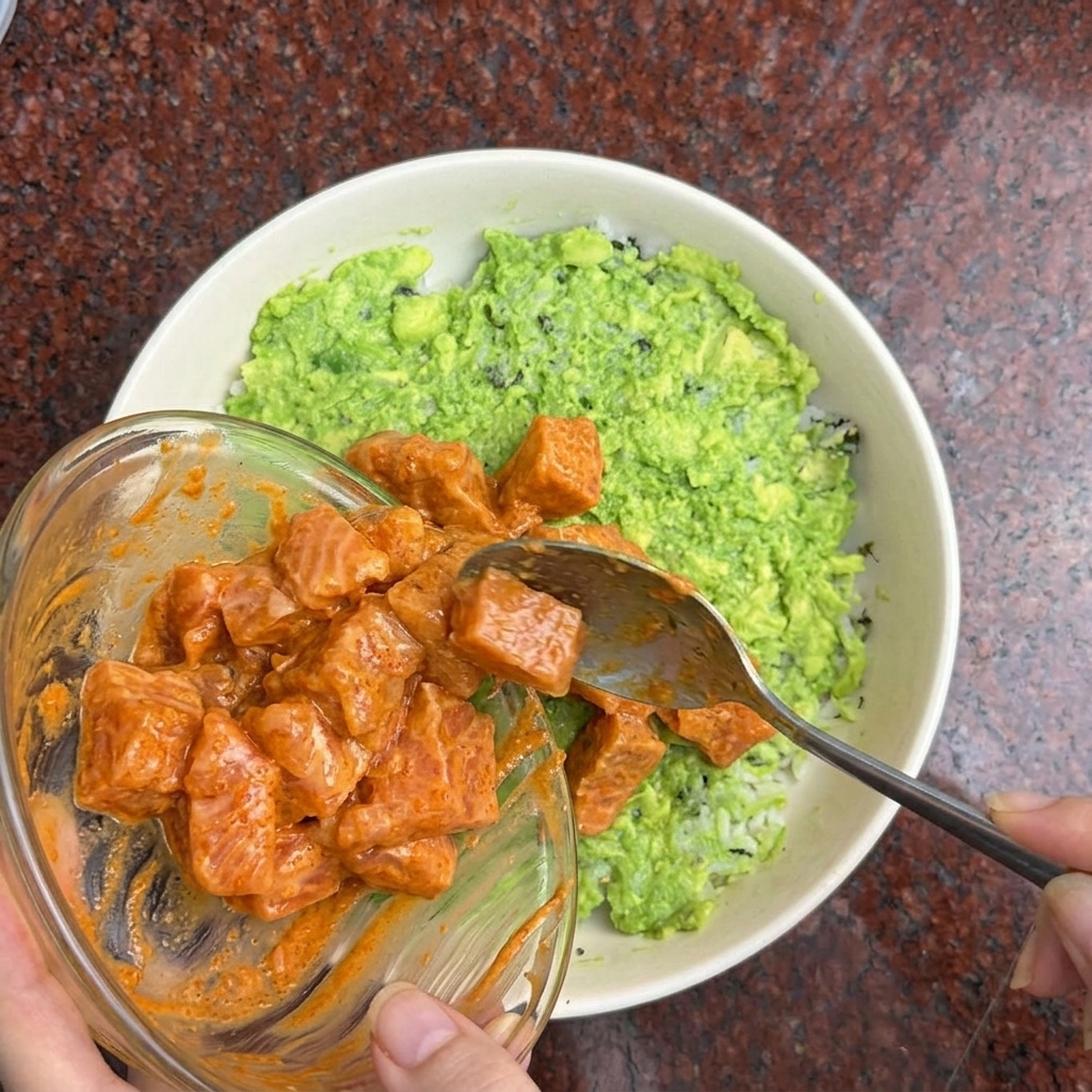 Dłoń nakładająca łyżką kostki łososia w pomarańczowej marynacie na miskę z ryżem i awokado.