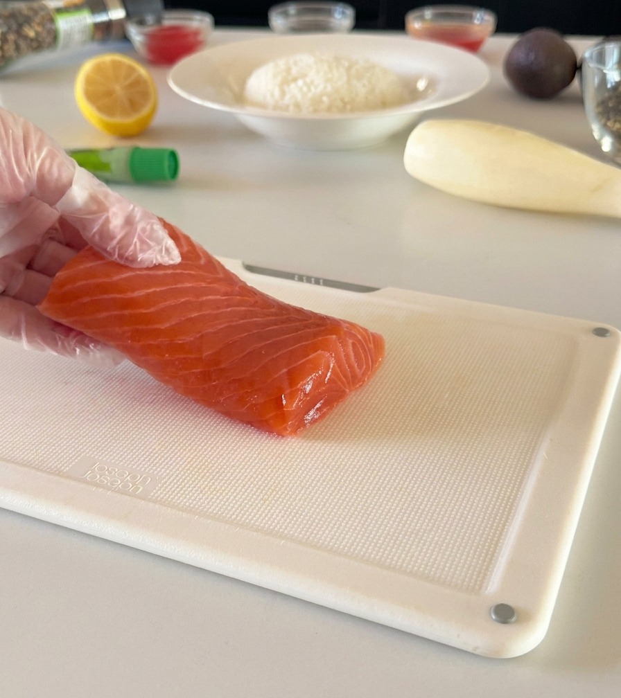 Dłoń w rękawiczce trzymająca świeży filet z łososia na białej desce, przygotowana do krojenia w kostkę.