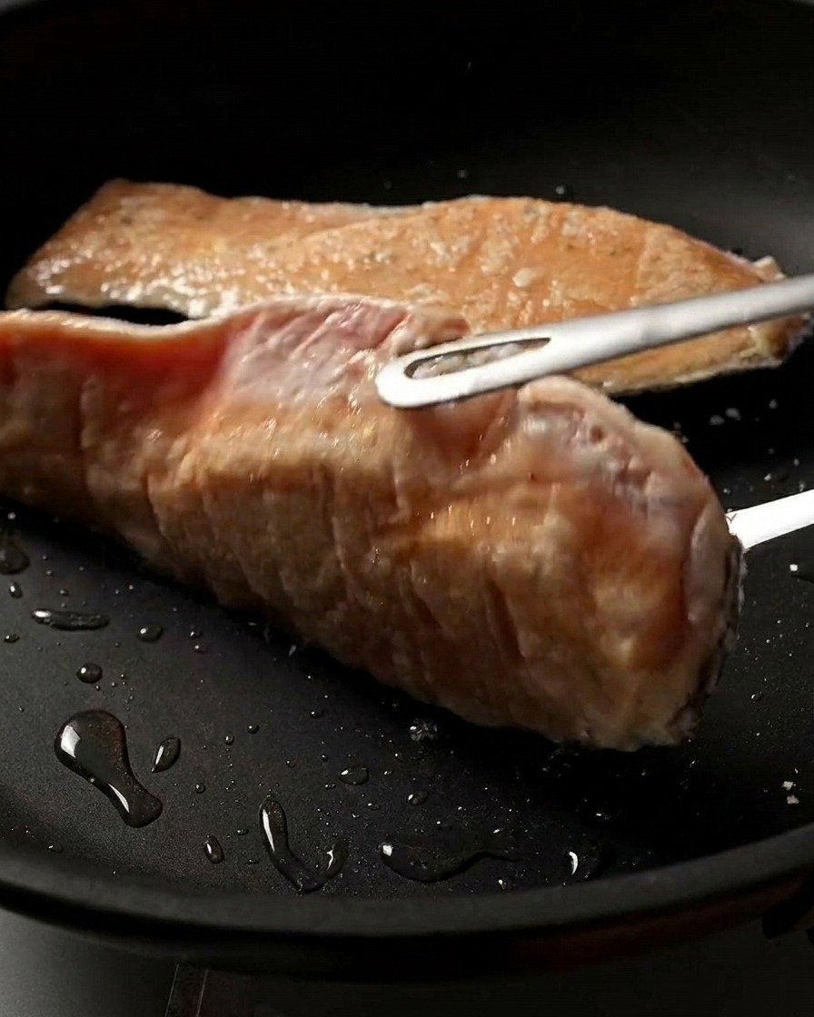 Zbliżenie na filet z łososia obracany na patelni, pokazujące jego idealnie zarumienioną i chrupiącą, usmażoną powierzchnię.