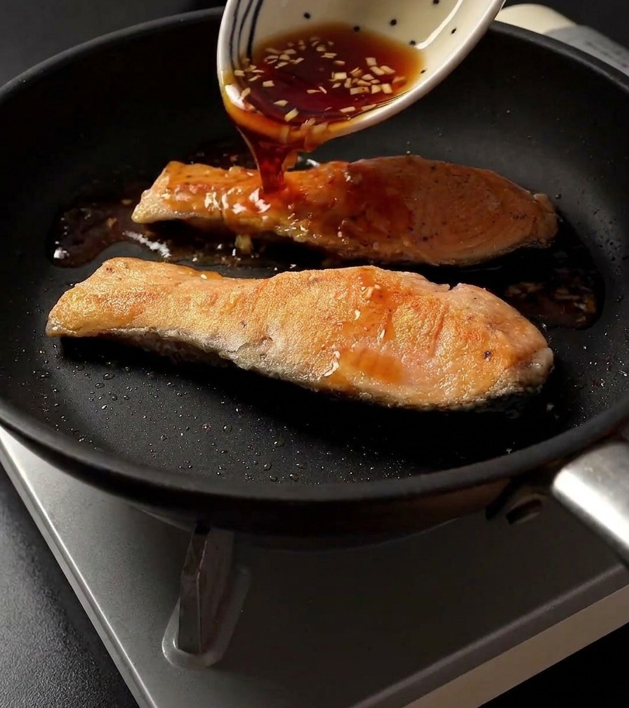Wlewanie ciemnego sosu teriyaki ze wzorzystej białej miski na dwa usmażone filety z łososia na patelni.