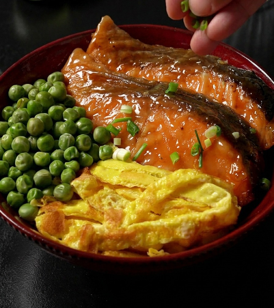 Dłoń posypująca posiekaną zieloną dymką gotową miskę ryżu zwieńczoną łososiem w glazurze teriyaki, paskami jajka i zielonym groszkiem.