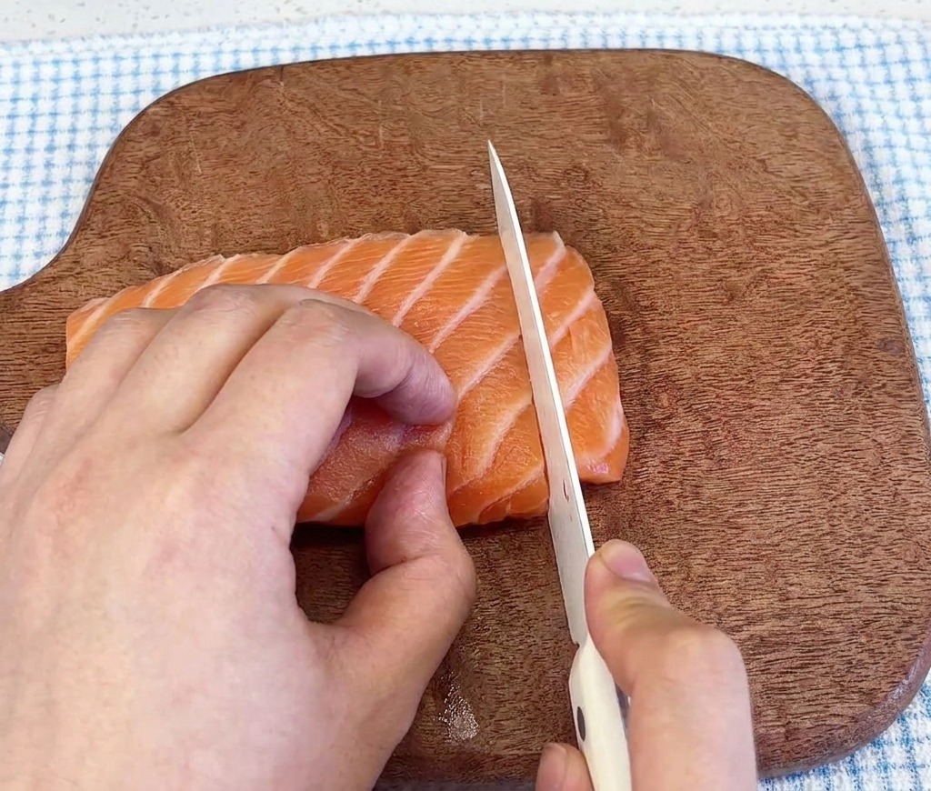 Zbliżenie na ostry nóż krojący świeży, marmurkowy filet z łososia na ciemnej drewnianej desce.