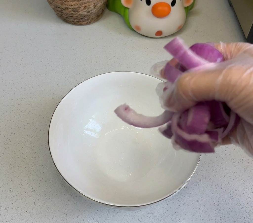 Osoba wrzucająca pokrojoną czerwoną cebulę do białej ceramicznej miski na czystym blacie kuchennym.