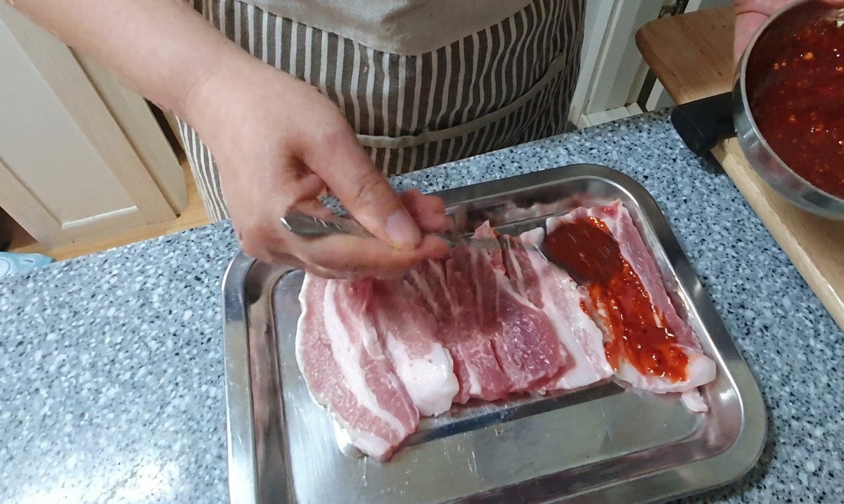 Dłoń nakładająca łyżką gęstą, intensywnie czerwoną marynatę chili na surowe plastry boczku ułożone na srebrnej tacy.