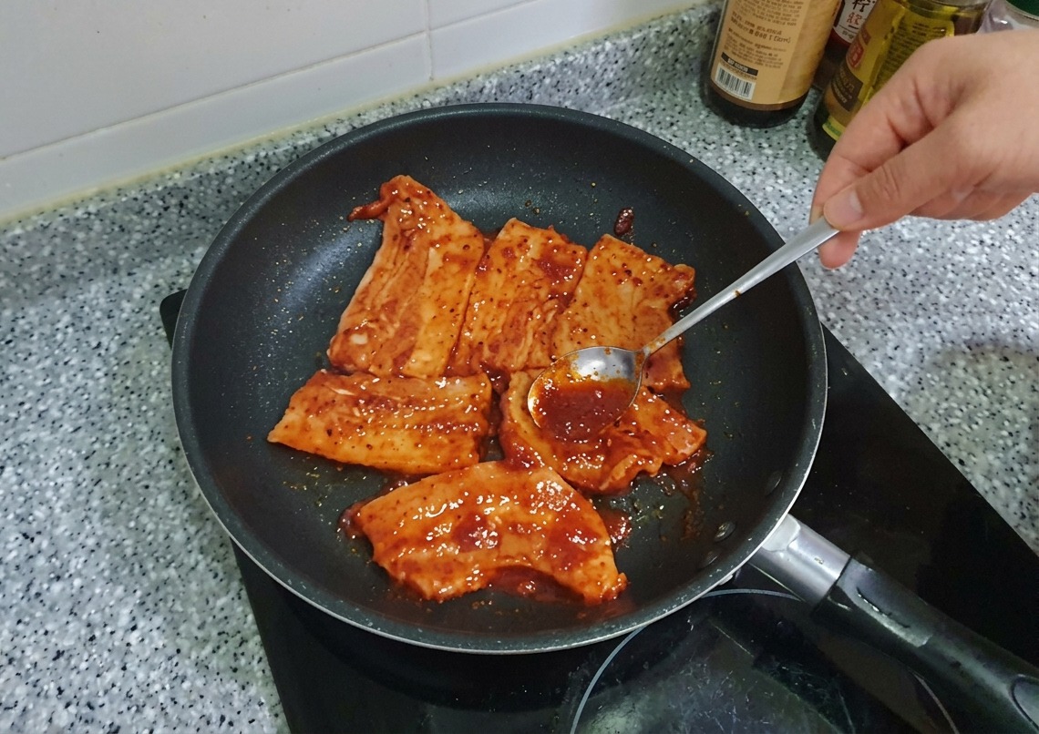 Łyżka rozprowadzająca gęstą, czerwoną marynatę Gochujang na plastrach boczku na patelni.