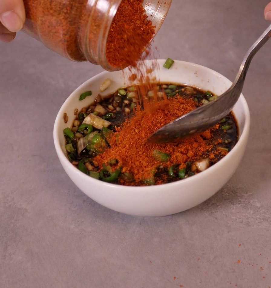 Jasnoczerwona drobna papryka chili w proszku wsypywana ze słoika do miski z przygotowaną bazą sosu i zielonym chili.