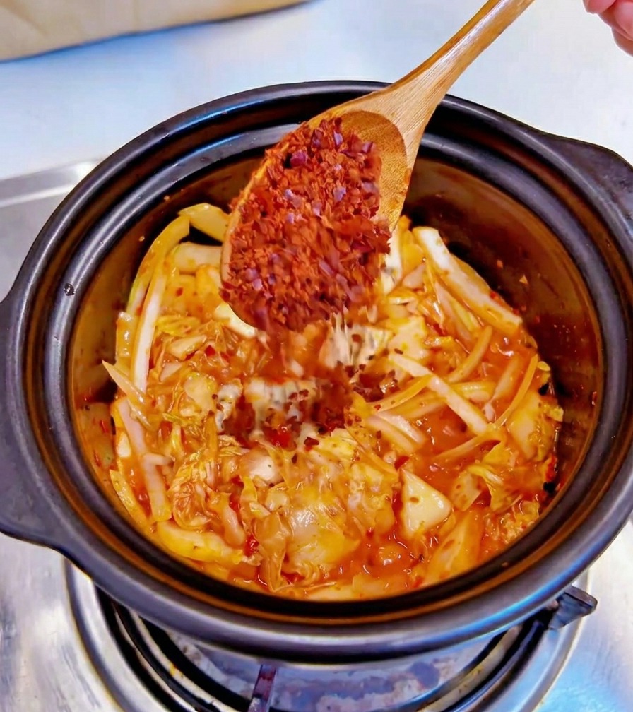 Drewniana łyżka wsypująca grubo mielone płatki chili do czarnego garnka z kimchi i cebulą.