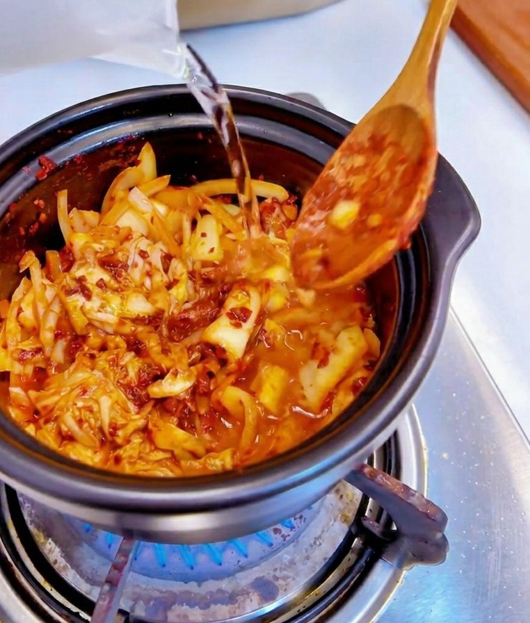 Woda wlewana z przezroczystej miarki do czarnego garnka z pikantnym kimchi i cebulą.