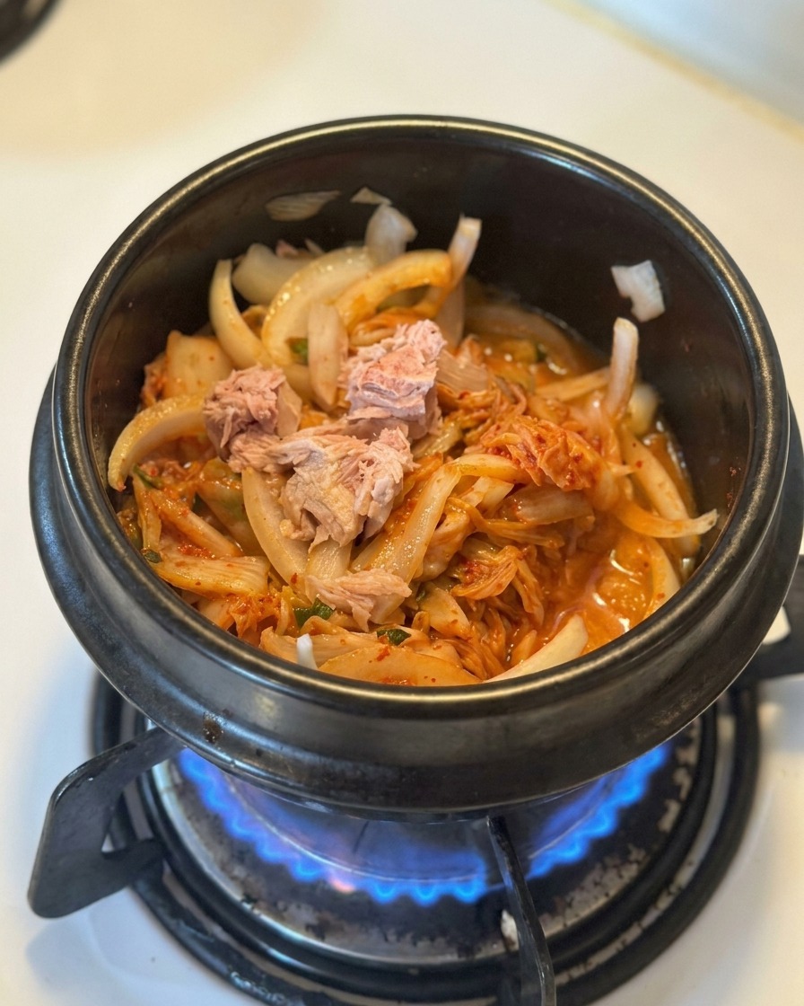 Kawałki tuńczyka z puszki ułożone na pierzynce z podsmażonego kimchi i cebuli w czarnym garnku.