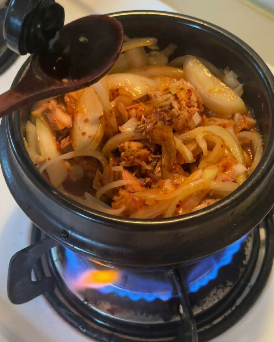 Drewniana łyżka wlewająca ciemny sos sojowy do czarnego glinianego garnka z podsmażoną cebulą, tuńczykiem i kimchi.