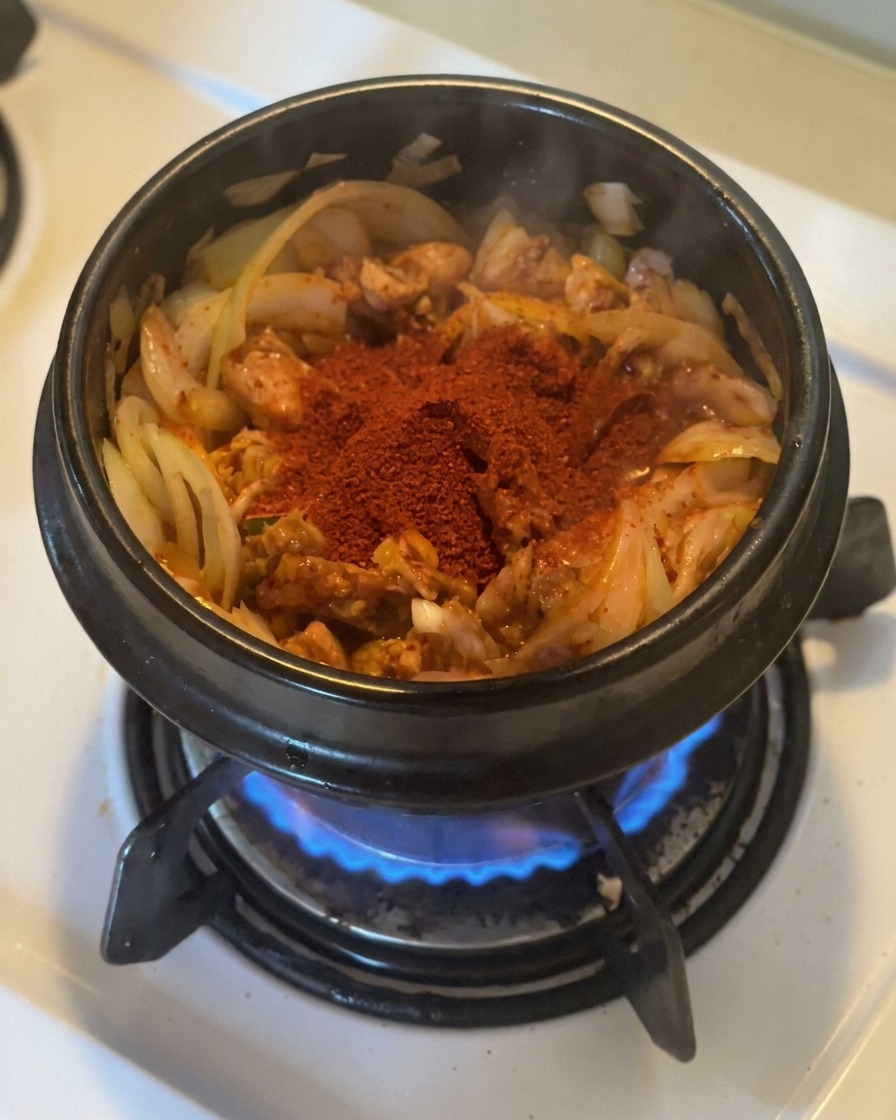Duża porcja jasnoczerwonego gochugaru na podsmażonym kimchi z cebulą w czarnym kamiennym garnku nad płomieniem gazowym.