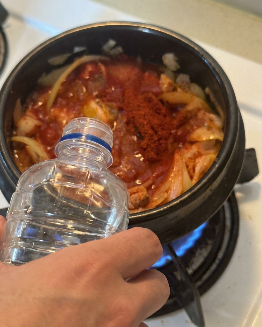 Woda wlewana z przezroczystej plastikowej butelki do garnka wypełnionego doprawionym na czerwono kimchi i tuńczykiem.
