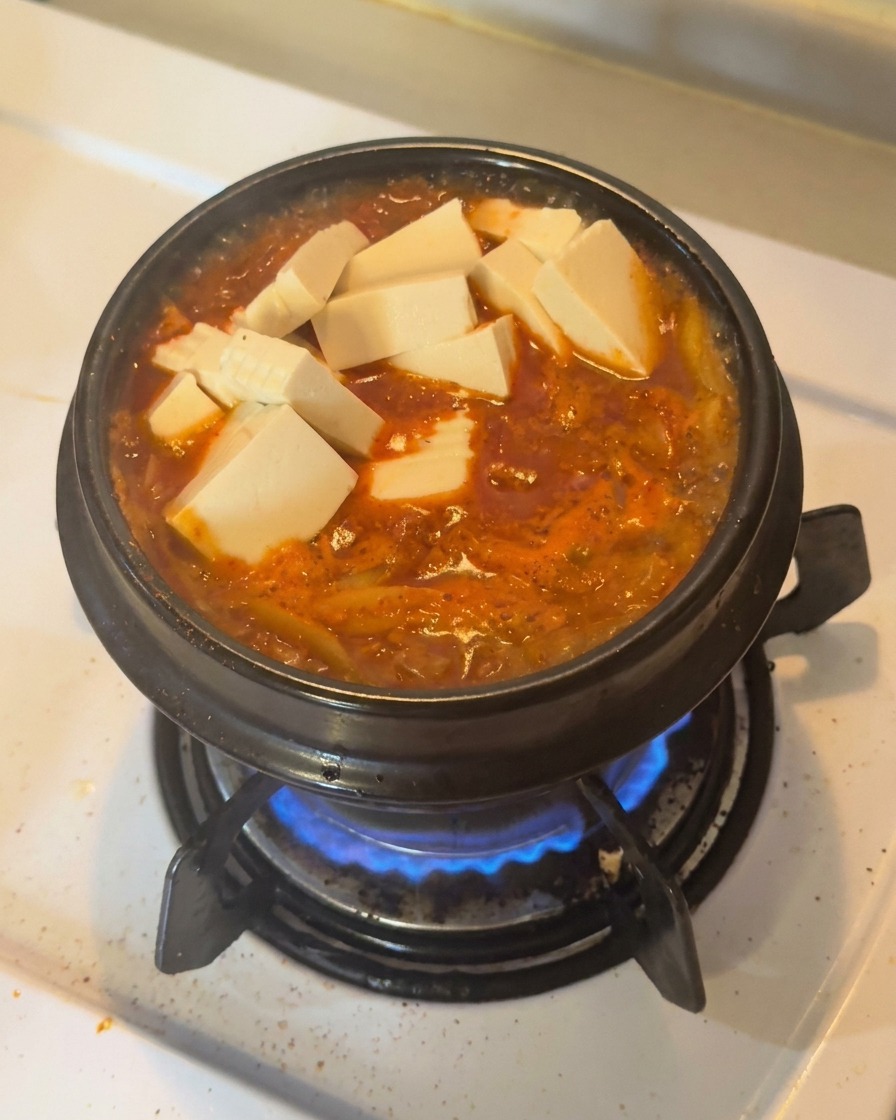 Kostki świeżego białego tofu i plasterki zielonej papryczki chili ułożone na wierzchu bulgoczącego, czerwonego gulaszu kimchi w czarnym glinianym garnku.