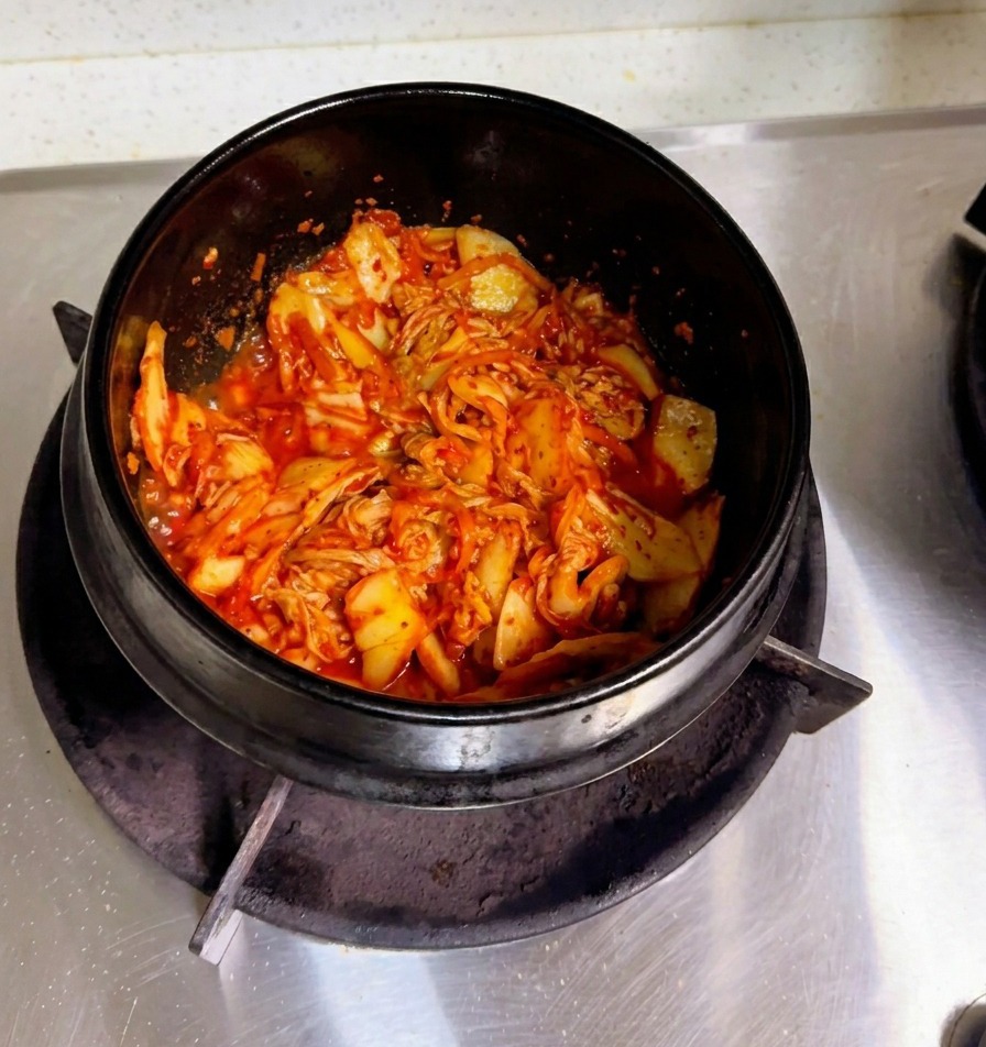 Dłoń wlewająca mętną wodę ryżową do kamiennego garnka z podsmażonym kimchi.