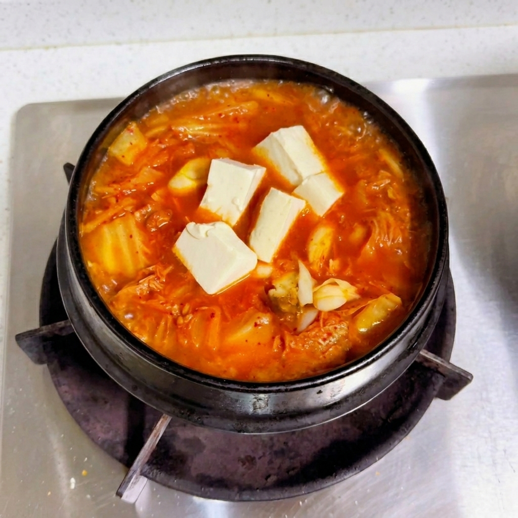 Kostki świeżego białego tofu ułożone na wierzchu bulgoczącego, czerwonego bulionu kimchi w czarnym garnku.